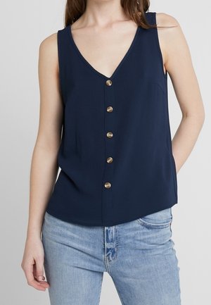 Woman wearing a sleeveless navy blue V-neck button-up top paired with light blue jeans, standing with one hand in pocket.