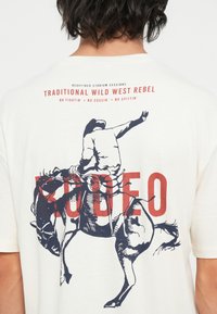 Off-white majica s grafikom rodeo jahača u mornarsko plavoj boji s crvenim natpisom "RODEO". Materijal je mekan s klasičnim dizajnom okruglog izreza.