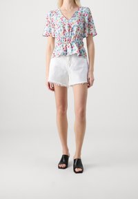 Blusa a fantasia floreale con vita arricciata, maniche corte e scollo a V, abbinata a pantaloni corti bianchi sfilacciati e sandali neri senza tacco.