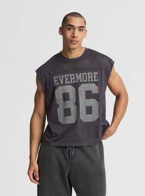 Giovane uomo con capelli corti che indossa una canotta nera in mesh con la scritta "EVERMORE 86" sulla parte anteriore e pantaloni della tuta scuri.