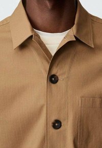 Chemise marron clair en tissu lisse, avec un col pointu, des manches courtes, deux boutons foncés et une poche poitrine.