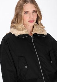 Schwarze Bomberjacke mit einem flauschigen beigen Kragen, Reißverschluss, und zwei Fronttaschen mit Druckknöpfen. Strukturiertes Außenmaterial.