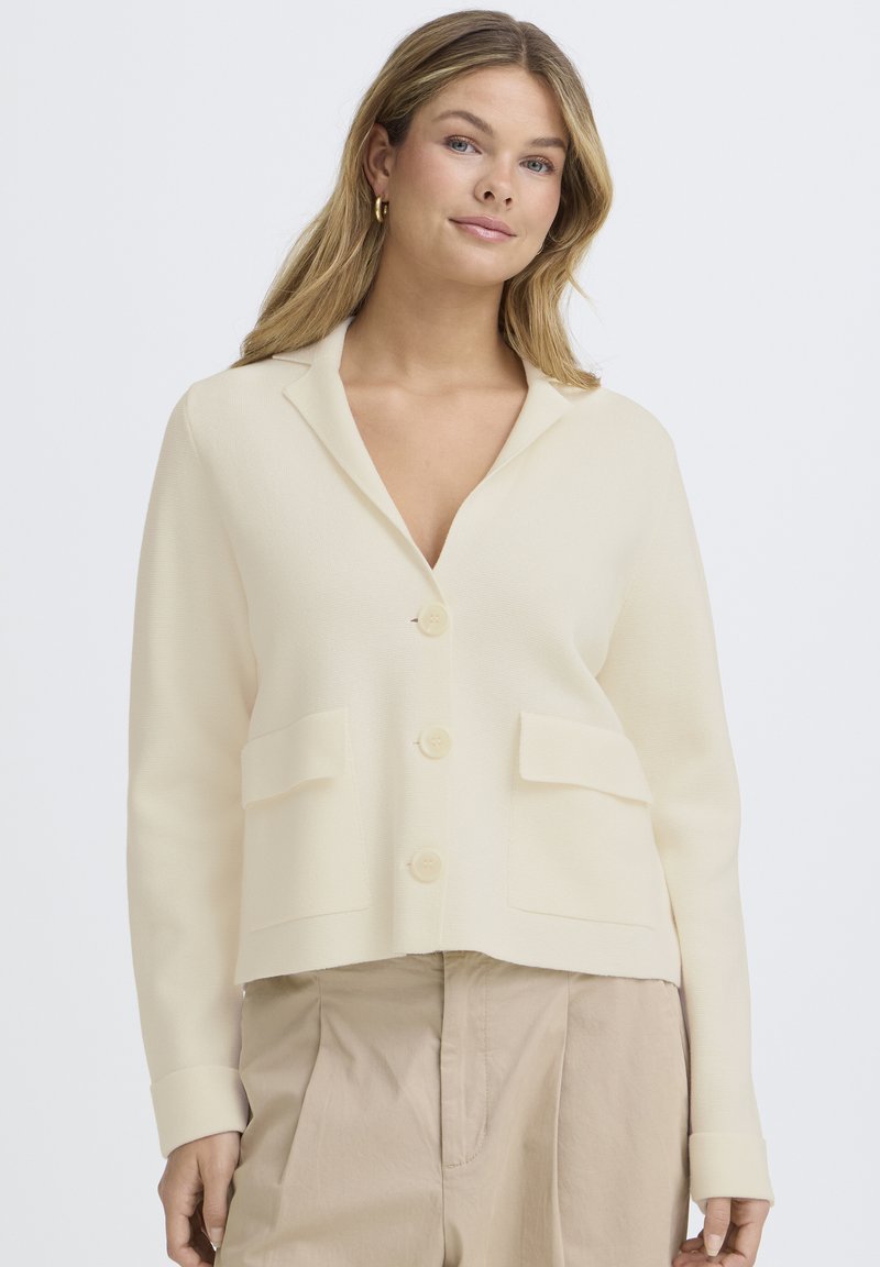 Femme aux longs cheveux blonds portant des boucles d'oreilles créoles dorées, un cardigan crème boutonné avec des poches, et un pantalon beige taille haute, debout devant un fond blanc.