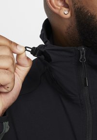 Chaqueta negra con un alto cuello y cremallera. El cuello tiene una cinta para ajustar. La tela parece suave con un acabado mate.
