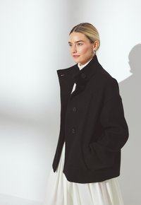Veste en laine noire avec une finition texturée, coupe oversize, col haut et boutons proéminents. Portée sur une robe blanche pour créer un contraste.