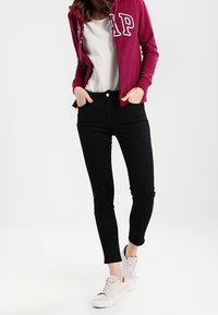 Sudadera con cremallera en color marrón con logo blanco, camiseta gris, jeans skinny negros y zapatillas ligeras. Conjunto casual con un diseño ajustado.