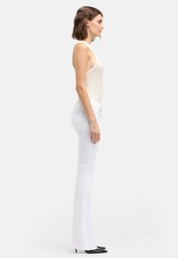 Jeans bianchi a zampa d'elefante con vestibilità slim, realizzati in denim, abbinati a un top smanicato beige chiaro e décolleté nere con punta a punta.