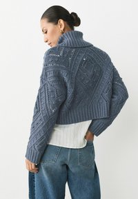 Kortad marinblå stickad tröja med intrikata mönster, ribbad krage och ärmslut, bärs över en vit strukturerad skjorta, i kombination med lösa blå jeans.