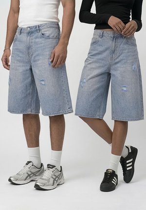 To personer iført lyseblå distressed denimshorts til knæet med hvide sokker og sneakers mod en ensfarvet baggrund.