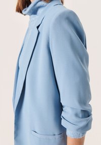Blazer azul claro con una tela suave, hombros estructurados, mangas enrolladas y un único bolsillo en la parte delantera. Cuello alto texturizado visible debajo.