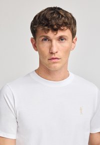 Vit kortärmad T-shirt i bomull med en liten beige logotyp på vänster bröst. Crewneck-design med en mjuk textur.