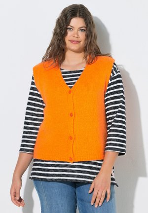 Donna con capelli castani mossi che indossa un gilet arancione brillante senza maniche con bottoni sopra una camicia a righe bianche e nere e jeans blu.