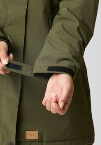 Olivengrøn jakke med en glat overflade, velcro lukning ved håndleddene og en tan farvet label med "Swedemount" ved kanten.