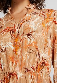 Robe fleurie en tissu doux et léger. Présente une base beige chaud avec des motifs abstraits orange et brun, un col à volants et une taille froncée.