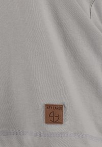 T-shirt in tessuto grigio chiaro con un'etichetta in pelle marrone recante il testo "KEY LARGO". I dettagli di cucitura all'orlo aggiungono una leggera texture.