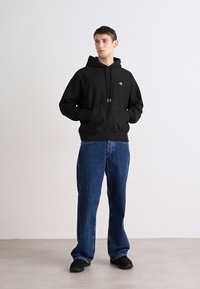 Čierna mikina s kapucňou a klokankou, v kombinácii so voľnými modrými džínsy. Model stojí na neutrálnom pozadí, má na sebe čierne topánky.