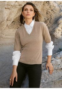 Beige kasjmiertrui met een V-hals en korte mouwen, gedragen over een wit overhemd met knopen. Het model draagt zwarte jeans en toont een ring aan haar hand.