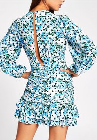 Žena v květinových a puntíkovaných šatech s nabráním, s nafouklými dlouhými rukávy a otevřenými zády, ruce opřené o boky.