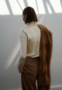 Svetlo bež turtleneck majica z visoko ovratnikom, v kombinaciji z mehkimi rjavimi hlačami in puhastim rjavim plaščem, ki je položeno čez eno ramo.