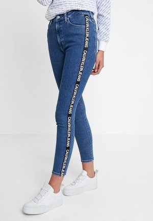 Jean slim bleu avec bande latérale noire comportant le texte blanc répété « Calvin Klein Jeans », associé à des baskets blanches et un haut à motifs.