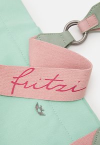 Helles grünes Canvas-Täschchen mit pinkem Stoffgurt, der den Markennamen "fritzi" in geschwungener Schrift zeigt, silberne Flügel-Logodetails und Metallbeschläge.
