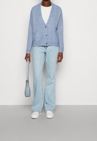 Hellblauer Strickcardigan mit V-Ausschnitt, drei Knöpfen und zwei Fronttaschen, kombiniert mit hellblauen ausgestellten Jeans und weißen Turnschuhen.