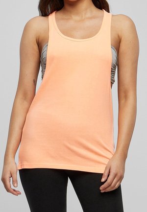 Kobieta w brzoskwiniowej, bezrękawnikowej koszulce na ramiączkach na szarym staniku sportowym i czarnych legginsach, stojąca na jednolitym tle.