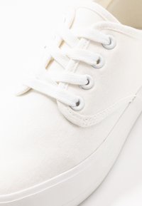 Sneaker bianca in tela con sei passanti, lacci bianchi e una superficie texturizzata. Sola in gomma bianca spessa per una maggiore stabilità.