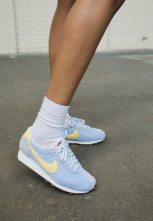Lichtblauwe Nike sneakers met gele accenten, voorzien van een gestructureerd bovenwerk, witte veters en een rubberen zool. Draagt witte enkelsokken.