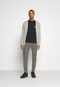 Siva zip-up hoodie, crna majica s džepom, maslinaste vojne hlače i crne tenisice. Moderni casual stil s praktičnim detaljima.