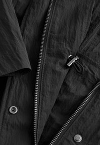 Sort krøllet stofjakke med to sølv lynlåse, trykknapper og en sort snor med metal lås.