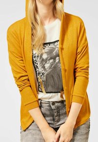 Cardigan à capuche jaune en tissu léger, doté d'une fermeture à boutons et de poignets côtelés, porté par-dessus un t-shirt à imprimé.