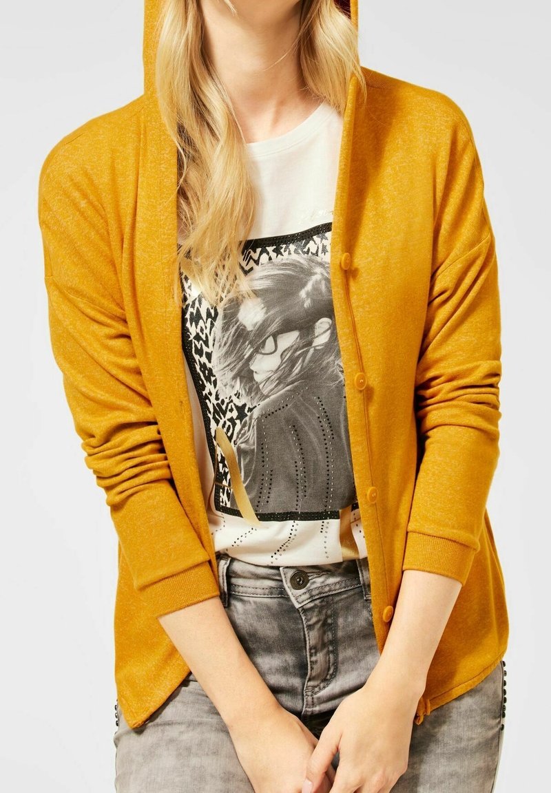 Cardigan à capuche jaune en tissu léger, doté d'une fermeture à boutons et de poignets côtelés, porté par-dessus un t-shirt à imprimé.