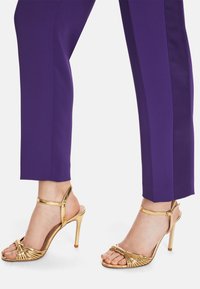 Gold strappy high-heeled sandals with a textured design. Paired with fitted purple trousers, highlighting sleek lines and a modern silhouette.