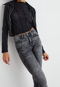 Chaqueta negra corta con cremallera plateada, combinada con una blusa negra texturizada y jeans ajustados grises descoloridos con detalles en los bolsillos.