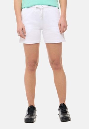 Personne portant un short blanc à cordon, un haut vert menthe et des chaussures de sport noires, debout les pieds écartés sur un fond uni.