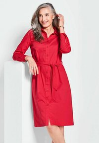 Rotes Knopfkleid mit gebundenem Taillengürtel, langen Ärmeln und einem Schlitz vorn. Glatte Textur, klassischer Kragen und taillierte Akzente.