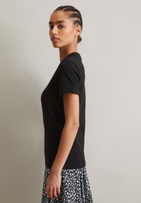 Schwarzes, eng anliegendes T-Shirt mit kurzen Ärmeln und geripptem Textur. Kombiniert mit einem gemusterten Rock mit weißen und blauen Motiven, Modell im Profil.