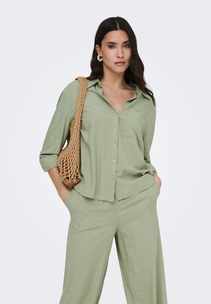 Femme aux longs cheveux foncés portant une chemise boutonnée vert clair et un pantalon assorti, portant un sac d'épaule tressé beige.