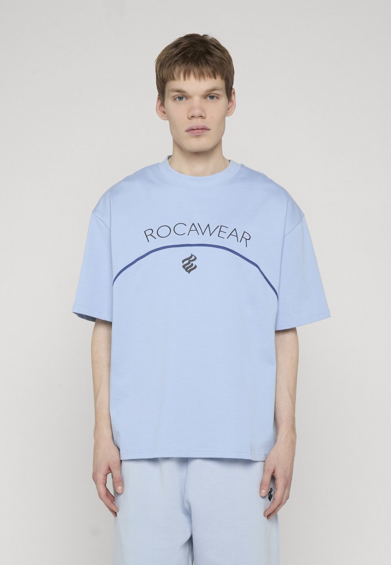 Jeune homme aux cheveux courts portant un T-shirt Rocawear bleu clair et un short assorti, debout devant un fond neutre uni.