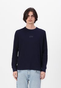 Chemise à manches longues bleu marine avec un col rond et une texture côtelée, arborant un logo sombre subtil sur la poitrine. Portée avec un jean bleu clair.