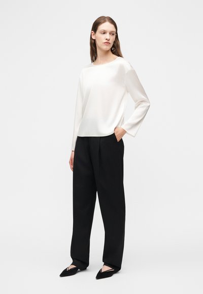 Γυναίκα που στέκεται με το χέρι στην τσέπη, φοράει ένα άνετο λευκό πουκάμισο με μακριά μανίκια, μαύρο παντελόνι με φαρδιά μπατζάκια και μαύρα μυτερά φλατ παπούτσια.