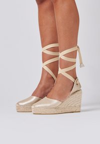 Espadrilles à semelles en jute tressé, en métal doré, avec des brides en tissu croisées autour des chevilles. Surface lisse et talons ouverts.