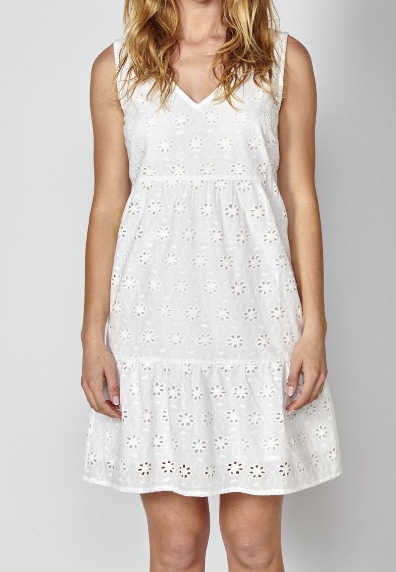 Robe blanche sans manches avec broderie florale ajourée, encolure en V et ourlet à volants froncés portée par une personne debout aux cheveux lâchés.