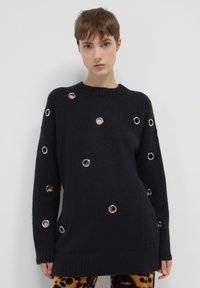 Maglione nero spesso a maglia con grandi occhielli argentati sparsi, scollo a girocollo e polsini a costine. Vestibilità oversize con un tessuto testurizzato.