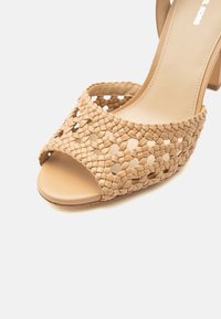 Μπεζ πέδιλο με ανοιχτά δάχτυλα και τακούνι, με πλεγμένα λουριά από δέρμα που σχηματίζουν ένα λουλουδένιο σχέδιο και μια λεία σόλα.