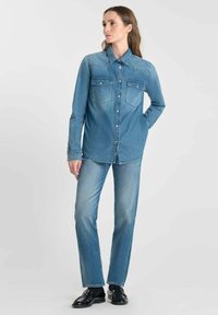 Kvinna iklädd en knappad denimskjorta med bröstfickor, ljusblå jeans och svarta loafers, stående med en hand bakom ryggen.
