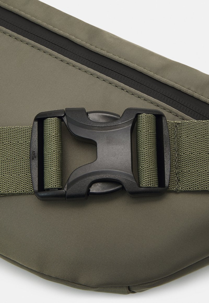 Bolsa de tela verde oliva con una cremallera negra, superficie texturizada, correa ajustable y un hebilla de plástico para un cierre seguro.