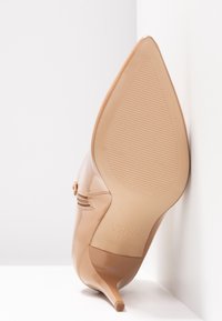 Chaussure à talon aiguilles beige en cuir verni à bout pointu, inclinée contre un mur blanc, montrant sa semelle texturée et son détail de fermeture éclair latérale.