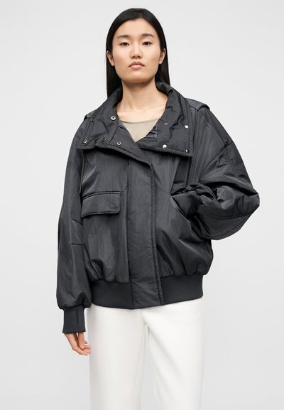 Juoda didelio dydžio striukė iš tekstūruotos medžiagos, su aukštu apykaklu, spaudėmis, priekiniu kišeniu ir ribomis rankogaliais.